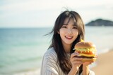 Surprise Asian Girl Holds And Eats Cheeseburger On By The Sea. Сoncept Surprise Asian Girl, Holding Eating Cheeseburger, Beautiful Beach Setting, The Power Of Advertising