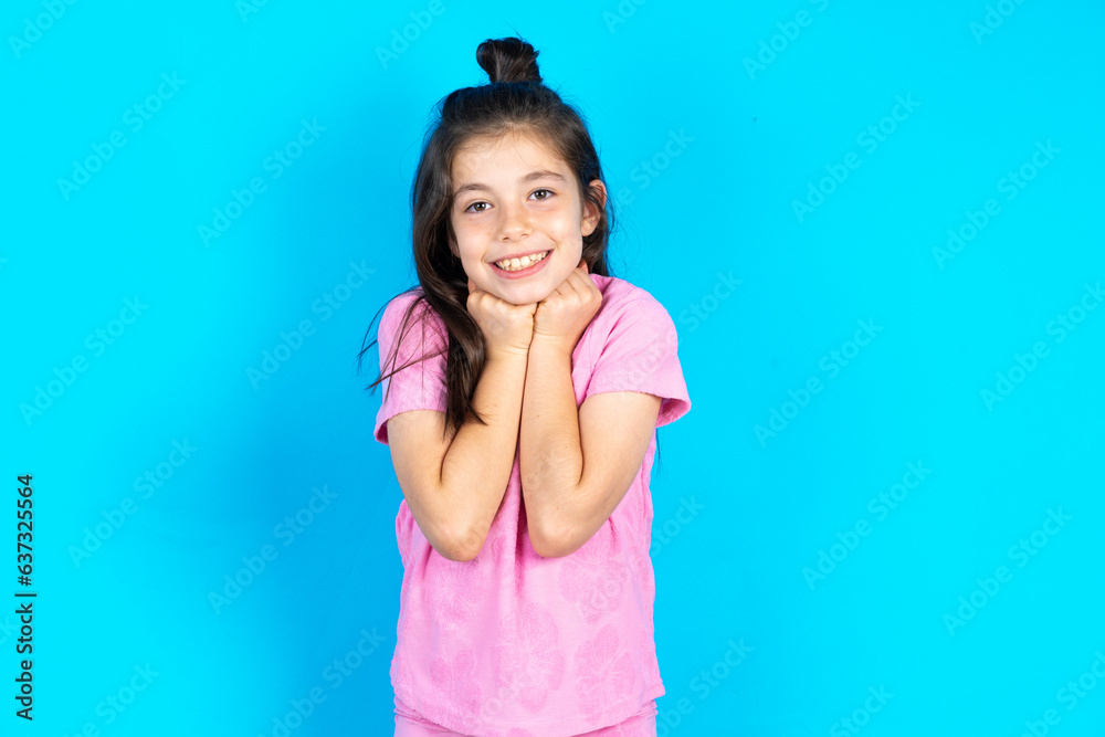 Satisfied Caucasian kid girl wearing pink t-shirt over blue background touches chin with both hands, smiles pleasantly, rejoices good day with lover Satisfied Caucasian kid girl wearing pink t-shirt over blue background touches chin with both hands, smiles pleasantly, rejoices good day with lover