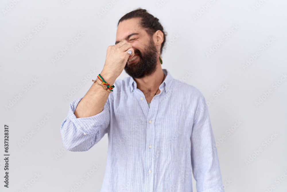 Hispanic man with beard wearing casual shirt smelling something stinky and disgusting, intolerable smell, holding breath with fingers on nose. bad smell Hispanic man with beard wearing casual shirt smelling something stinky and disgusting, intolerable smell, holding breath with fingers on nose. bad smell