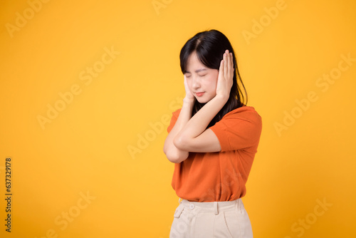 Tired asian woman 30s shouting in reaction to loud noise, covering her ear. Expression of stress and frustration isolated on yellow background.