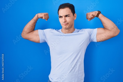 Young hispanic man standing over blue background showing arms muscles smiling proud. fitness concept.