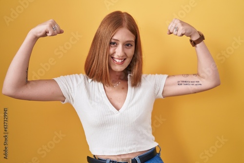 Young redhead woman standing over yellow background showing arms muscles smiling proud. fitness concept.