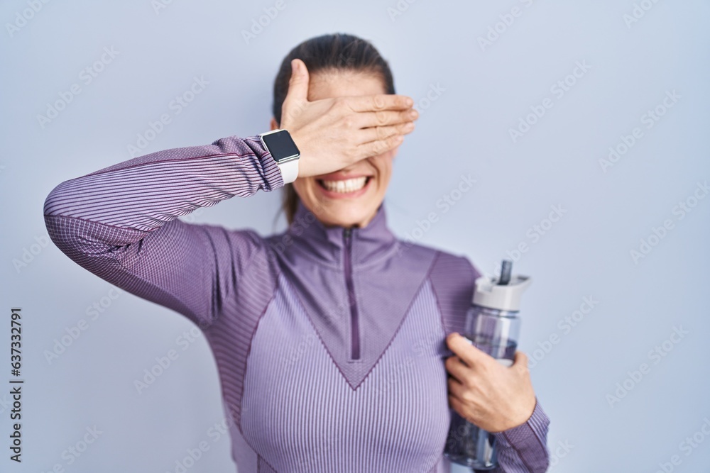 Beautiful woman wearing sportswear holding water bottle smiling and laughing with hand on face covering eyes for surprise. blind concept.