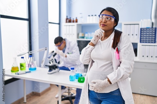 Young hispanic woman expecting a baby working at scientist laboratory serious face thinking about question with hand on chin  thoughtful about confusing idea