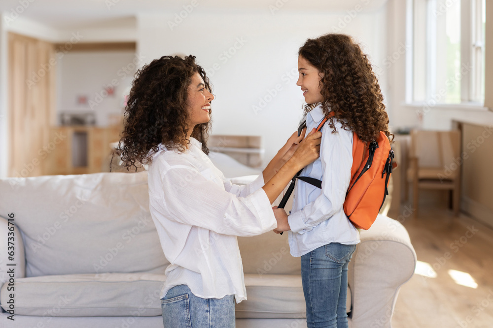 Back to school. Happy latin mother preparing schoolgirl daughter for first school day, helping to put on bag at home interior, side view. Mother and child bond concept - obrazy, fototapety, plakaty 