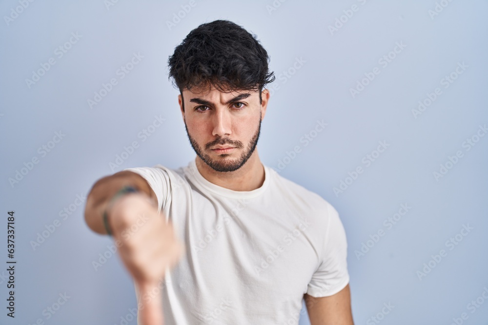 Hispanic man with beard standing over white background looking unhappy and angry showing rejection and negative with thumbs down gesture. bad expression. Hispanic man with beard standing over white background looking unhappy and angry showing rejection and negative with thumbs down gesture. bad expression.
