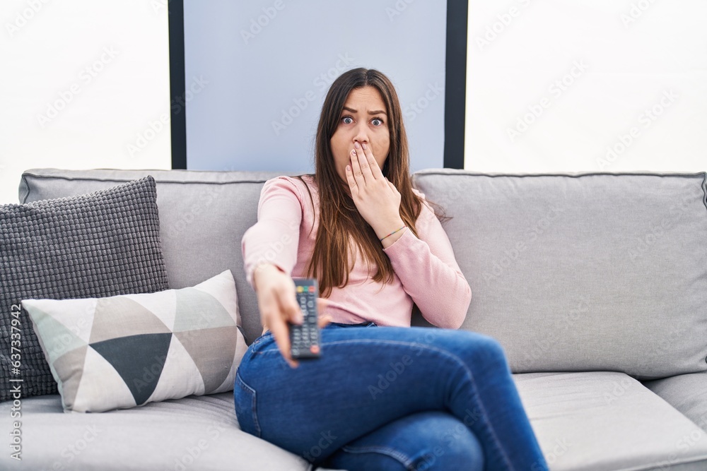 Young brunette woman holding television remote control covering mouth with hand, shocked and afraid for mistake. surprised expression Young brunette woman holding television remote control covering mouth with hand, shocked and afraid for mistake. surprised expression