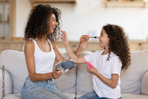 Happy latin mother and little daughter sitting on sofa at home, putting makeup to each other, having fun together, trying different beauty products