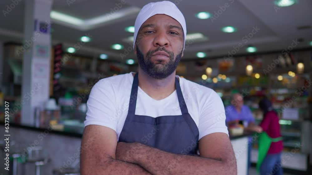 One young serious Black employee with arms crossed standing inside cafeteria front business One young serious Black employee with arms crossed standing inside cafeteria front business