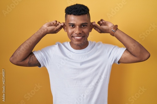 Young hispanic man standing over yellow background smiling pulling ears with fingers, funny gesture. audition problem