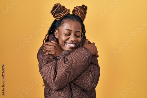 African woman with braided hair standing over yellow background hugging oneself happy and positive, smiling confident. self love and self care © Krakenimages.com