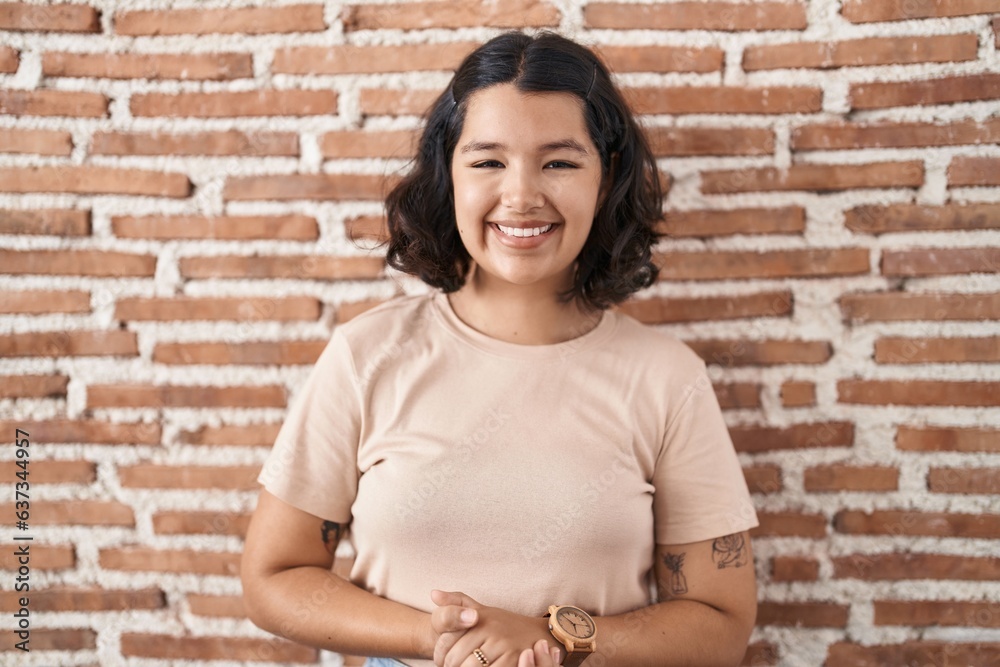 Young hispanic woman standing over bricks wall with hands together and crossed fingers smiling relaxed and cheerful. success and optimistic