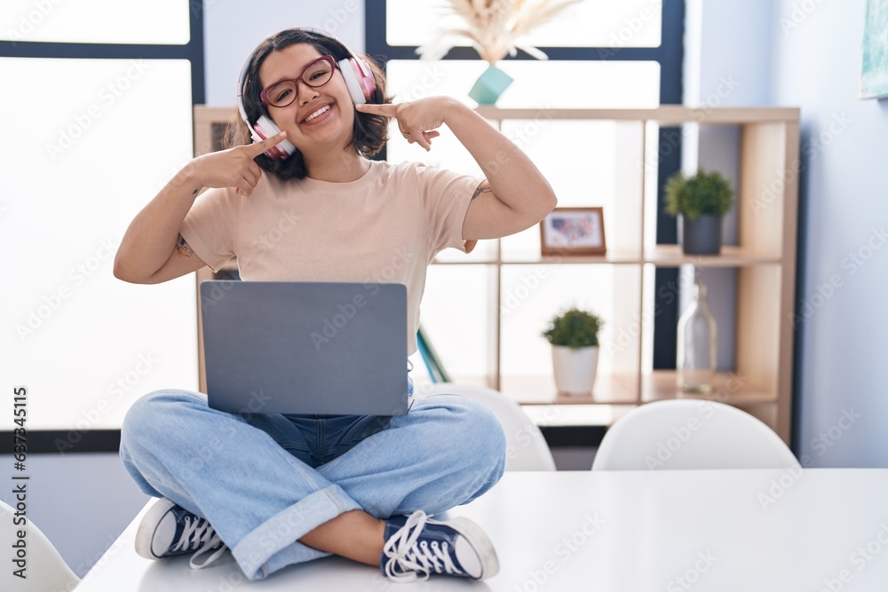 Young hispanic woman using laptop sitting on the table wearing headphones smiling cheerful showing and pointing with fingers teeth and mouth. dental health concept. Young hispanic woman using laptop sitting on the table wearing headphones smiling cheerful showing and pointing with fingers teeth and mouth. dental health concept.