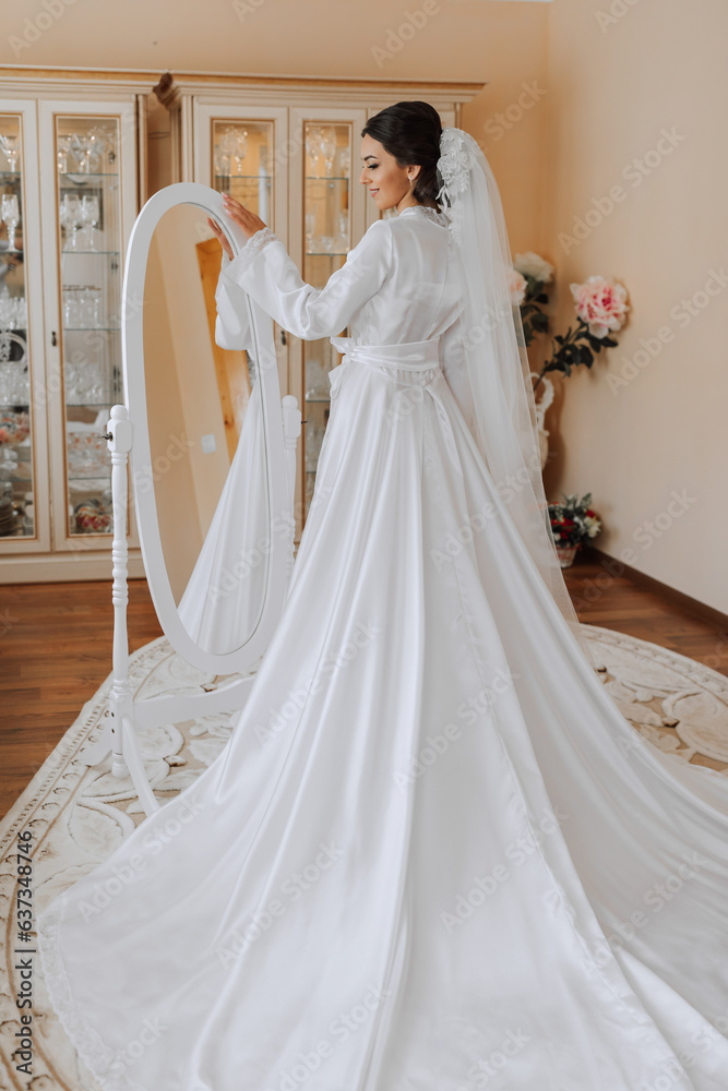 Fototapeta premium Happy dark-haired brunette bride in a satin robe and professional make-up standing in her room near the mirror and getting ready for her wedding day
