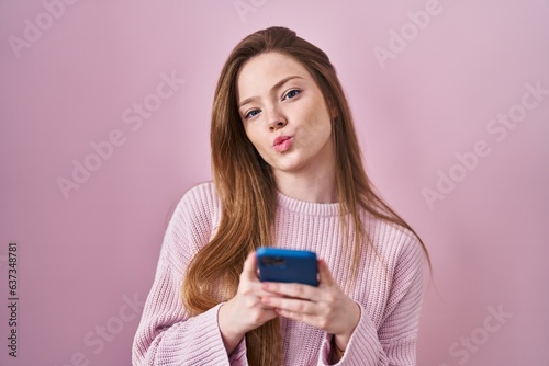 Young caucasian woman using smartphone typing message looking at the camera blowing a kiss being lovely and sexy. love expression.