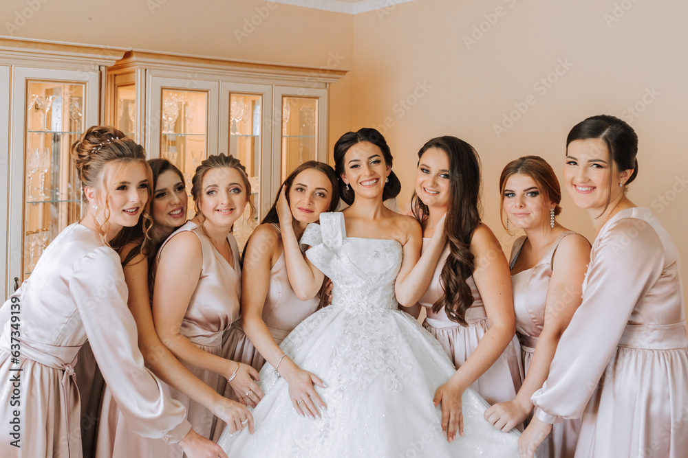 Stylish beautiful bridesmaids in matching silk dresses helping gorgeous brunette bride in white dress get ready for wedding, morning preparations, woman putting on dress