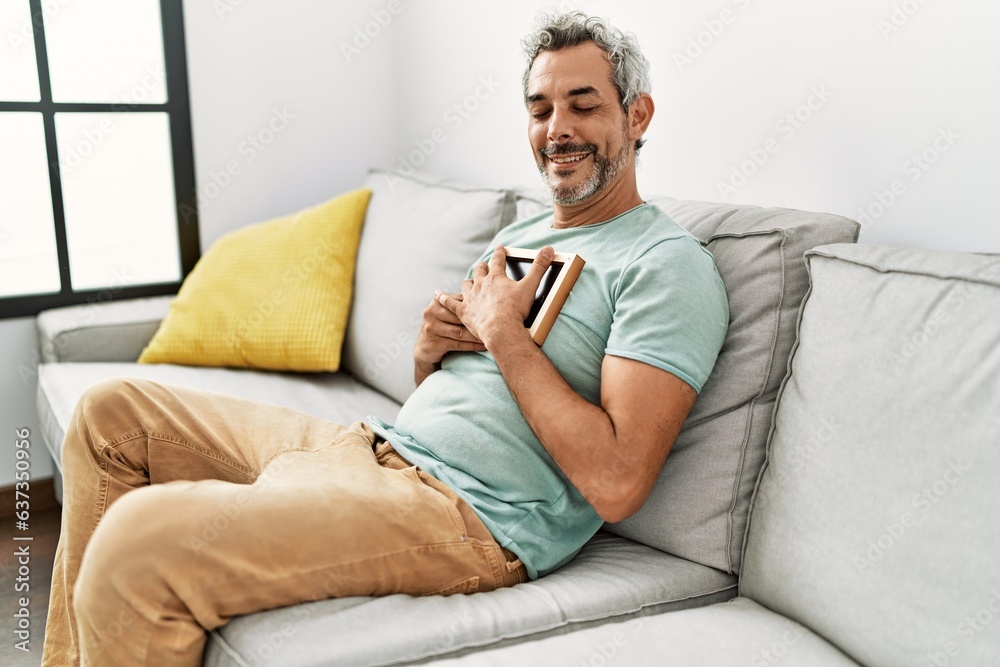 Middle age grey-haired man hugging picture sitting on sofa at home Middle age grey-haired man hugging picture sitting on sofa at home
