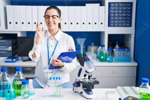 Young brunette woman working at scientist laboratory gesturing finger crossed smiling with hope and eyes closed. luck and superstitious concept.