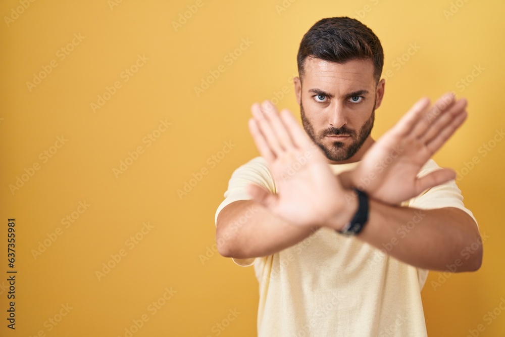 Handsome hispanic man standing over yellow background rejection expression crossing arms and palms doing negative sign, angry face Handsome hispanic man standing over yellow background rejection expression crossing arms and palms doing negative sign, angry face