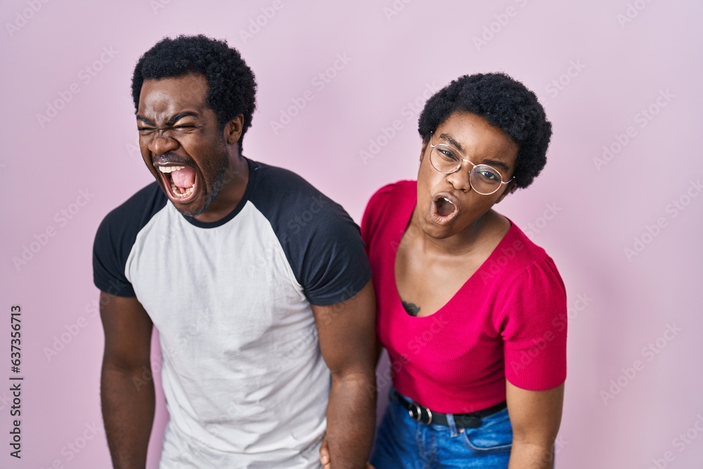 Young african american couple standing over pink background angry and mad screaming frustrated and furious, shouting with anger. rage and aggressive concept. Young african american couple standing over pink background angry and mad screaming frustrated and furious, shouting with anger. rage and aggressive concept.