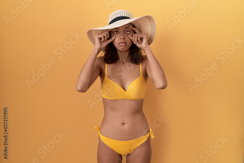 Young hispanic woman wearing bikini and summer hat trying to open eyes with fingers, sleepy and tired for morning fatigue