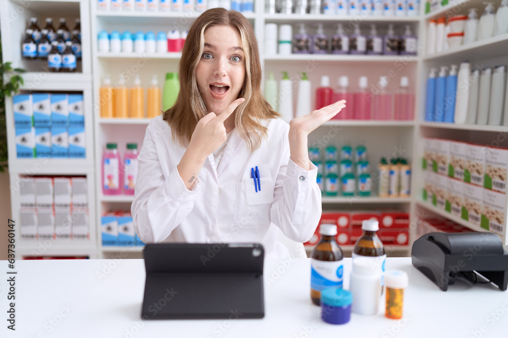 Young caucasian woman working at pharmacy drugstore using tablet amazed and smiling to the camera while presenting with hand and pointing with finger.