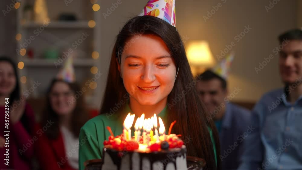 Young charming woman in party hat blowing out candles on birthday cake and looking at camera. Blurred group of friends rejoicing at background. Happy lady posing with dessert indoors Young charming woman in party hat blowing out candles on birthday cake and looking at camera. Blurred group of friends rejoicing at background. Happy lady posing with dessert indoors