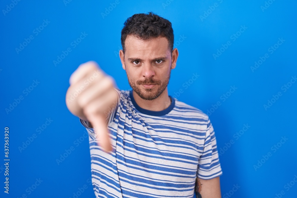 Young hispanic man standing over blue background looking unhappy and angry showing rejection and negative with thumbs down gesture. bad expression. Young hispanic man standing over blue background looking unhappy and angry showing rejection and negative with thumbs down gesture. bad expression.
