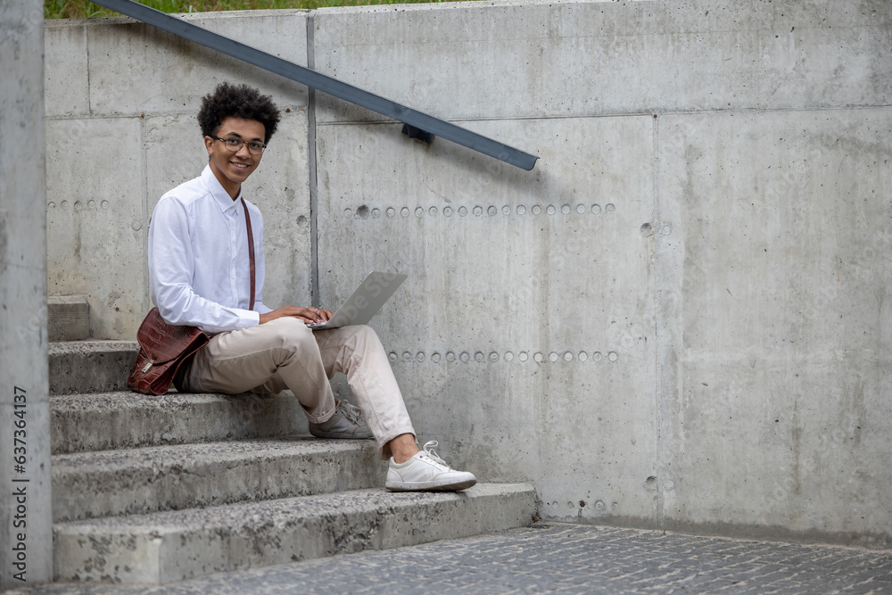 Curly-haired young man in white shirt sitting on the steps with laptop Curly-haired young man in white shirt sitting on the steps with laptop