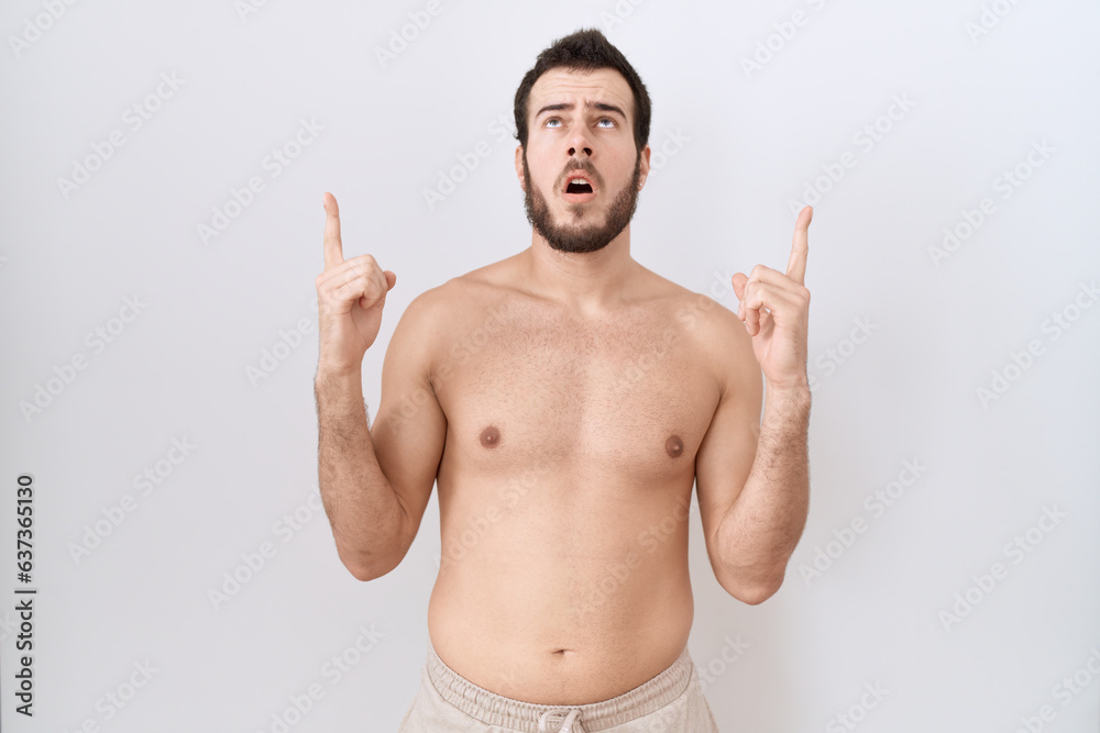 Young hispanic man standing shirtless over white background amazed and surprised looking up and pointing with fingers and raised arms.
