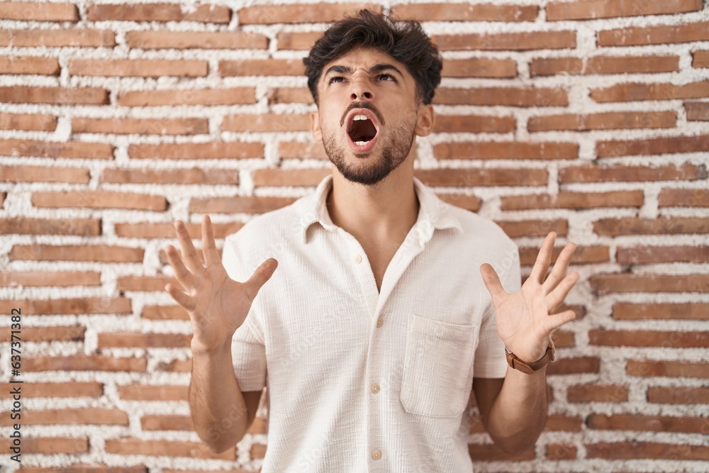 Arab man with beard standing over bricks wall background crazy and mad shouting and yelling with aggressive expression and arms raised. frustration concept. Arab man with beard standing over bricks wall background crazy and mad shouting and yelling with aggressive expression and arms raised. frustration concept.
