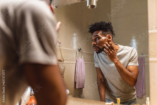 Handsome guy applying moisturizer and looking at himself while standing in front of the mirror in the bathroom.