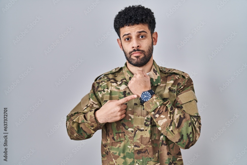Arab man wearing camouflage army uniform in hurry pointing to watch time, impatience, looking at the camera with relaxed expression
