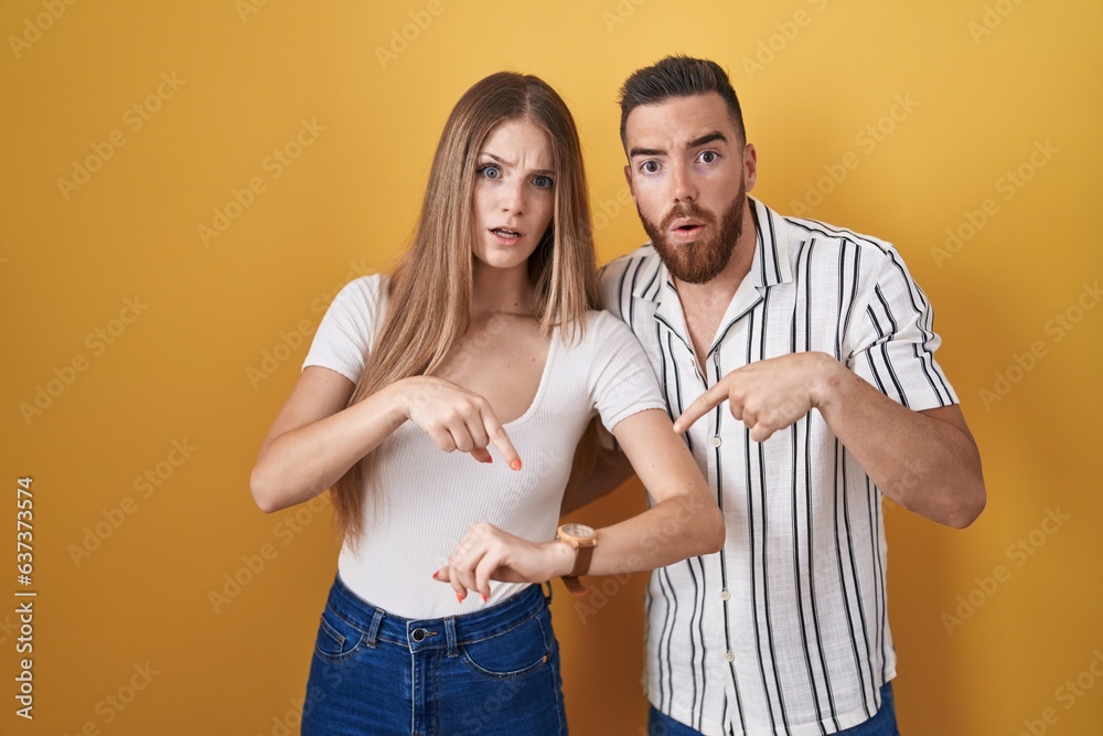 Fototapeta premium Young couple standing over yellow background in hurry pointing to watch time, impatience, upset and angry for deadline delay