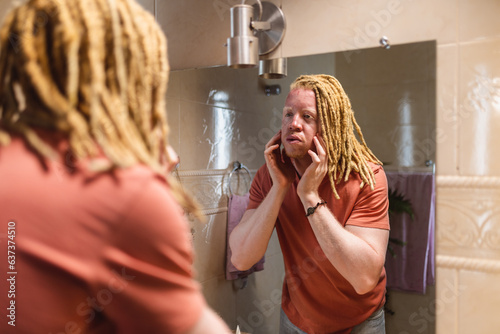 Homem albino estiloso com dreadlocks na frente do espelho do banheiro durante sua rotina de cuidados com a pele. photo