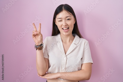 Chinese young woman standing over pink background smiling with happy face winking at the camera doing victory sign with fingers. number two. © Krakenimages.com