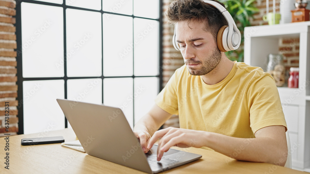 Fototapeta premium Young hispanic man using laptop and headphones sitting on table at dinning room