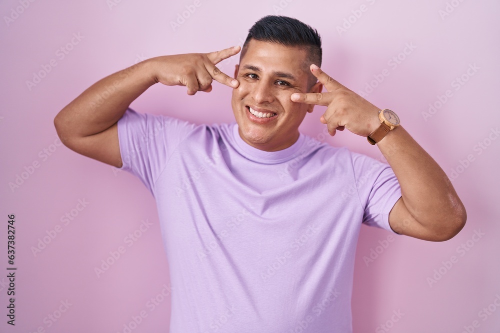 Young hispanic man standing over pink background doing peace symbol with fingers over face, smiling cheerful showing victory Young hispanic man standing over pink background doing peace symbol with fingers over face, smiling cheerful showing victory