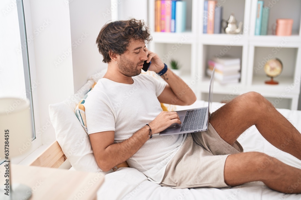 Young hispanic man using laptop talking on smartphone at bedroom
