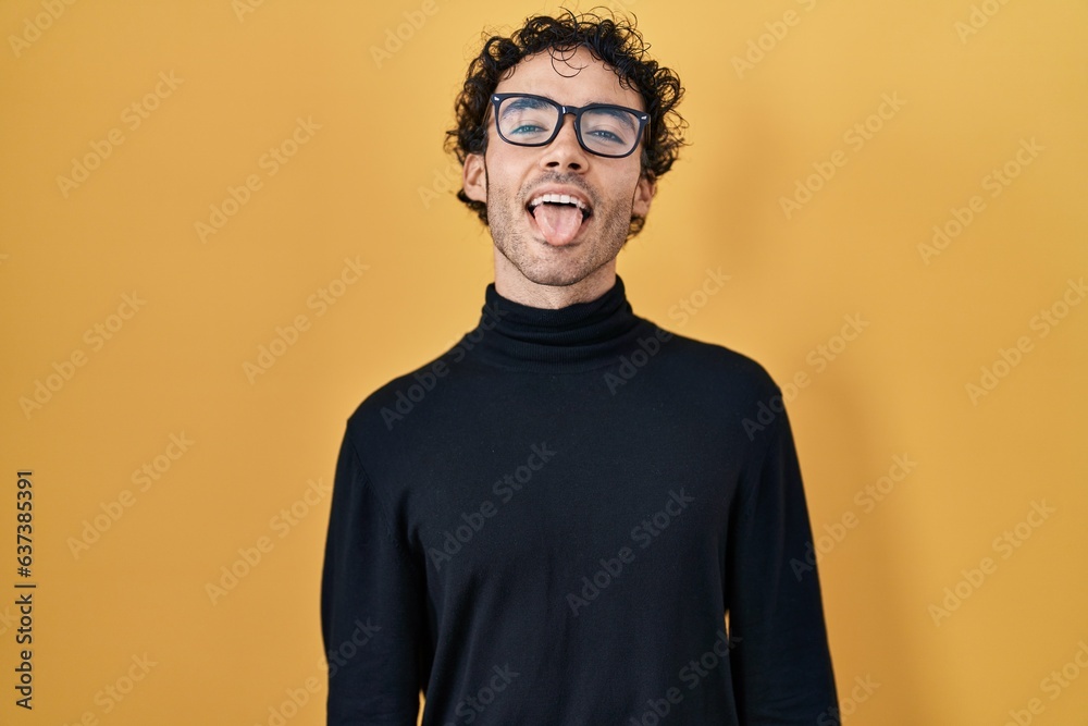 Hispanic man standing over yellow background sticking tongue out happy with funny expression. emotion concept. Hispanic man standing over yellow background sticking tongue out happy with funny expression. emotion concept.