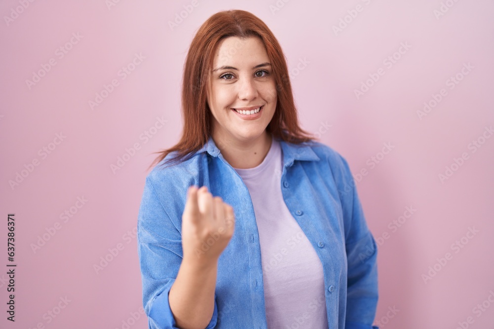 Young hispanic woman with red hair standing over pink background beckoning come here gesture with hand inviting welcoming happy and smiling Young hispanic woman with red hair standing over pink background beckoning come here gesture with hand inviting welcoming happy and smiling