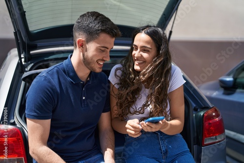 Young hispanic couple sitting on car trunk using smartphone at street