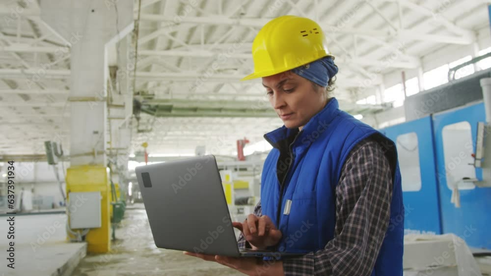 Medium shot of female technician in hardhat and workwear using laptop during workday at industrial factory Medium shot of female technician in hardhat and workwear using laptop during workday at industrial factory