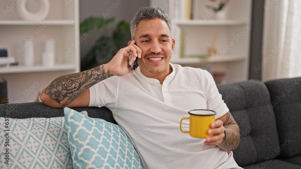 Young hispanic man talking on smartphone drinking coffee at home