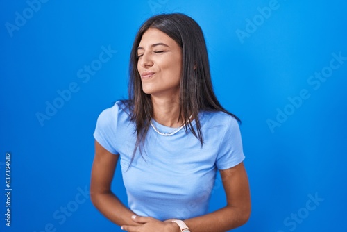 Brunette young woman standing over blue background with hand on stomach because indigestion, painful illness feeling unwell. ache concept.