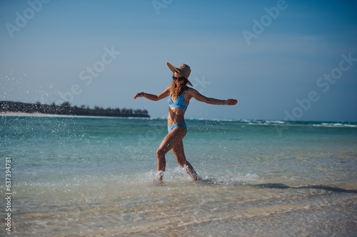 Portrait of a beautiful slim woman splashing water on the beach in a bikini and a straw hat.