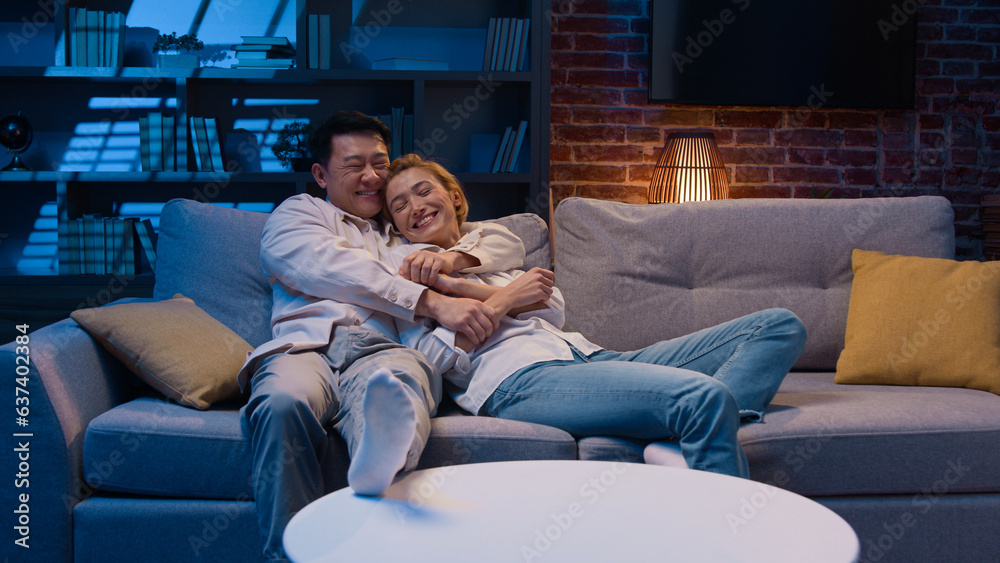 Happy multiracial diverse couple Asian man and Caucasian woman homeowners renters jumping on sofa in new apartment relax on couch at night evening laughing hugging real estate mortgage loan tenancy