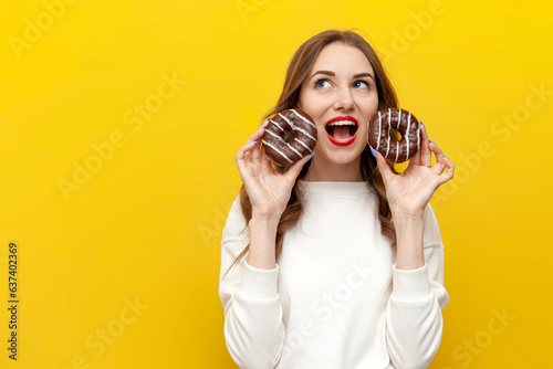 young cute girl holding two chocolate donuts over yellow isolated background, woman screaming with open mouth