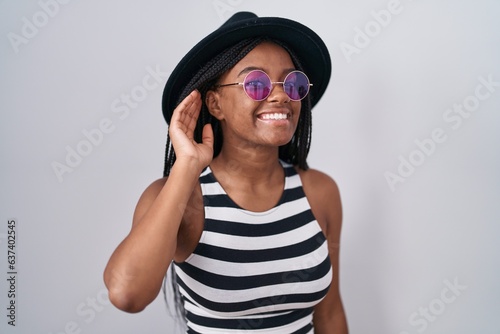 Young african american with braids wearing hat and sunglasses smiling with hand over ear listening an hearing to rumor or gossip. deafness concept.