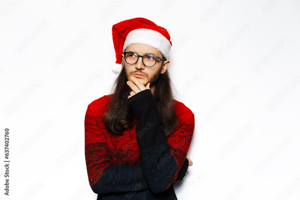 Studio portrait of young thoughtful man in red sweater, looking away, wearing Santa Claus hat and eyeglasses. Isolated on white background. Studio portrait of young thoughtful man in red sweater, looking away, wearing Santa Claus hat and eyeglasses. Isolated on white background.